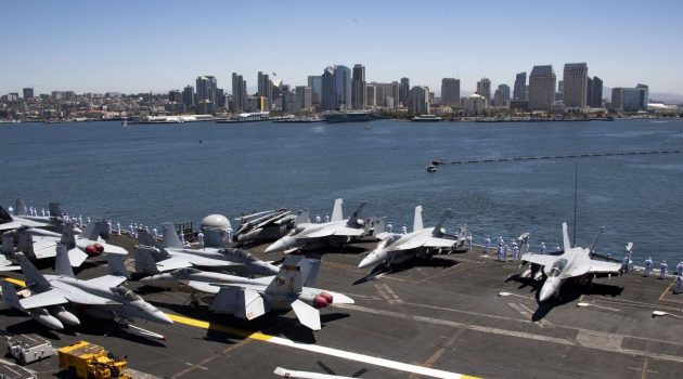 A real top gun with planes aboard an aircraft carrier and the San Diego skyline in the background.